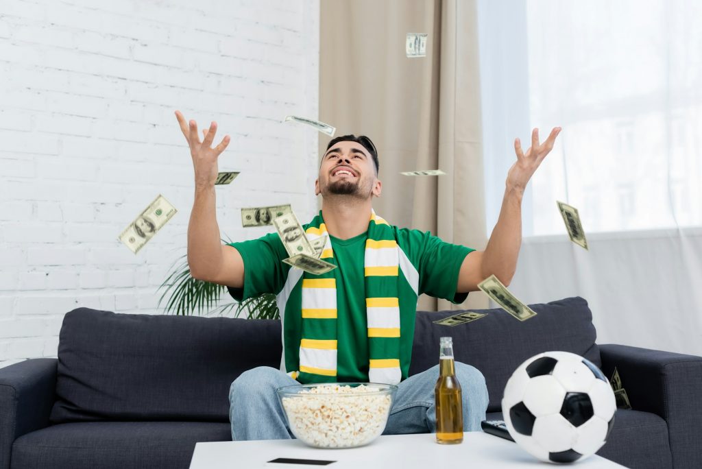 happy bookmaker throwing up dollar banknotes near soccer ball, beer and popcorn